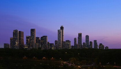 Naklejka premium A wide panoramic evening view of illuminated skyscrapers in the Toronto financial center rising above a dark treeline and highway during a purple twilight dusk in Ontario Canada