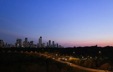 Naklejka premium A wide panoramic evening view of illuminated skyscrapers in the Toronto financial center rising above a dark treeline and highway during a purple twilight dusk in Ontario Canada