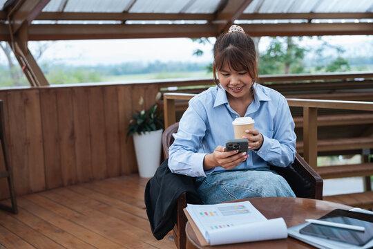 Young woman enjoys coffee while using smartphone in relaxed outdoor setting with financial documents on table, focusing on data analysis and planning discussion
