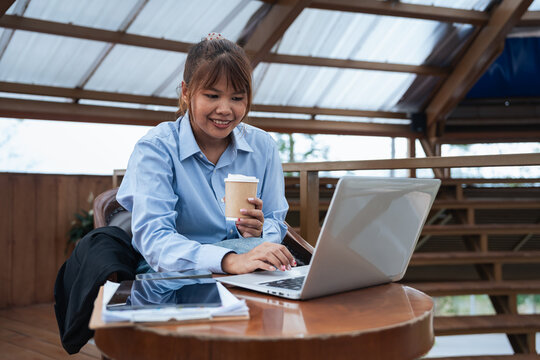 Young woman holding coffee cup working on laptop at wooden table with financial data and analysis papers planning business discussion in cozy outdoor workspace