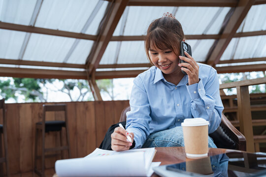 Young woman making phone call while doing financial planning and data analysis with documents and tablet on table during discussion in cozy cafe environment