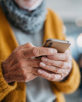 Hands grip phone with gentle focus. Senior wears warm yellow sweater. Soft scarf adds cozy contrast. Fingers tap screen, showing quiet attention