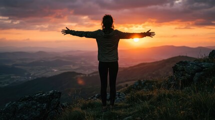 Fototapeta premium A silhouetted person stands on a rocky hilltop, arms outstretched towards a vibrant sunset. The individual faces the rising sun, seemingly embracing the expansive landscape before them.