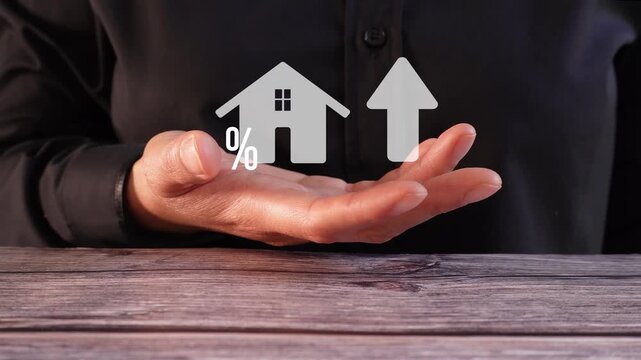 Close-up of a woman's hand at a table holding an animated house icon with a percentage sign, next to an upward-pointing arrow. Real Estate Topics