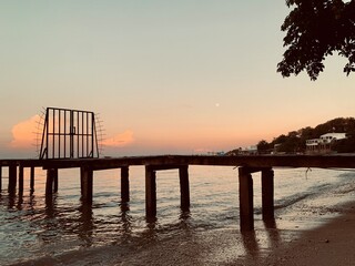 Obraz premium Weathered wooden pier extending over calm tropical waters at sunset. Warm evening tones, shoreline reflections, and dramatic silhouettes create a peaceful coastal travel background.