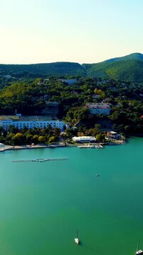 Vertical video. Abrau-Diurso, Krasnodar Krai, Russia. Lake Abrau, emerald water surrounded by forested hills and the historic winery village, Drone footage under bright summer sun. Drone footage. Ric
