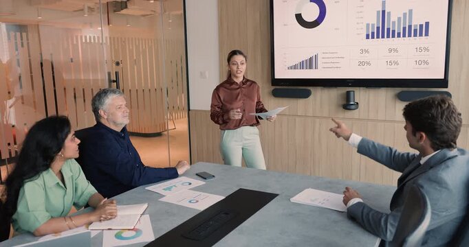 Confident young female business leader woman presenting sales and expenses report on large electronic whiteboard, pointing at graphic benchmarks on screen, speaking to diverse team