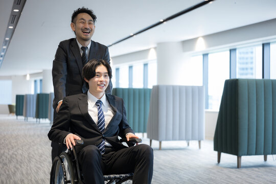 オフィスで車椅子利用者をサポートしながら移動するビジネスマンのバリアフリーの支援や障害者雇用などのイメージ全身