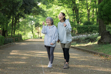 Obraz premium 新緑背景でシニア女性と娘世代の女性が公園でランニングやウォーキング(歩く,走る)のスポーツや運動を楽しむ健康的なイメージ全身