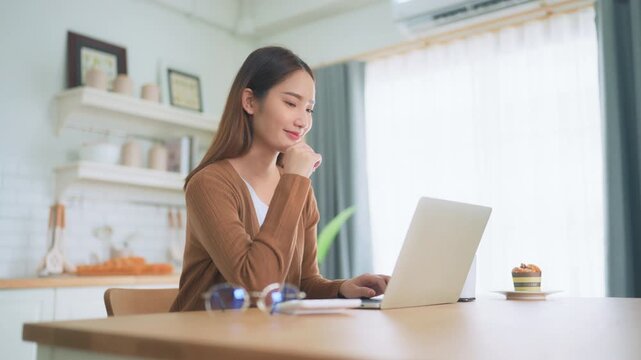 Beautiful young Asian woman working with laptop computer in home kitchen. Work at home