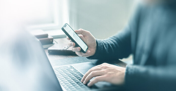 A person using a smartphone while typing on a laptop. The scene is bright and modern, suggesting a productive workspace environment.