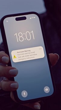 Vertical video. Woman Holding Smartphone With Hurricane Warning Notification Near Stormy Coastline at Dusk, Emergency Alert and Natural Disaster Preparedness