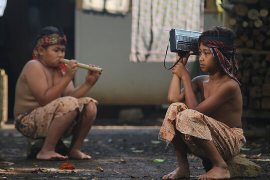 Two traditional Indonesian village boys sitting outdoors, one playing a bamboo flute while the other listens to a vintage portable transistor radio.