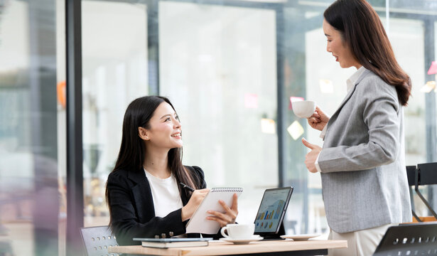 Two Asian businesswomen chatting during coffee break in office.