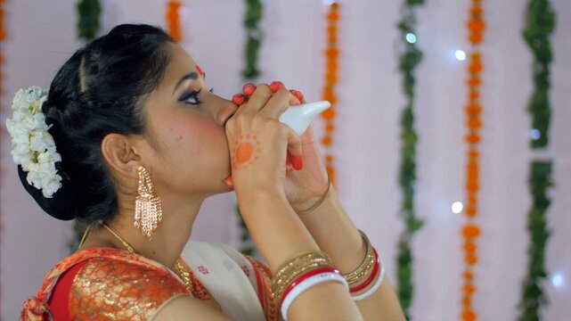 Bengali woman blowing a conch shell during the Durga Puja celebrations in India. Side view of a traditionally dressed Indian female blows conch shell (Shankh) at the Durga Puja - festival background