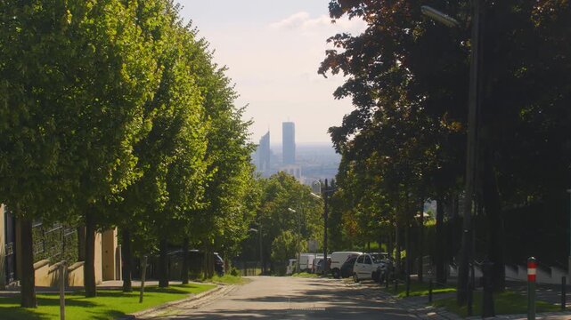 Narrow residential street with row of green trees and view of distant modern skyscrapers in Paris suburb.