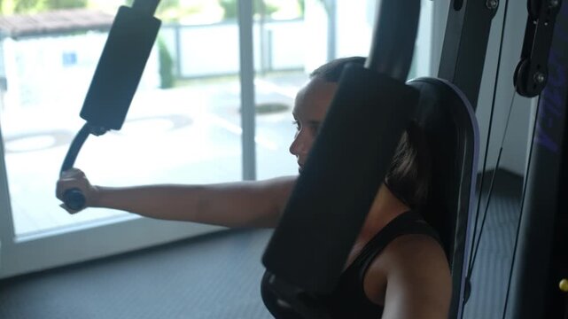 Close-up side view of young woman in black top performing chest press exercise on a gym machine with focused expression during training, slow motion. Fitness concept