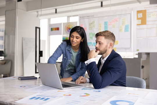 Young 30s Indian woman showing presentation on laptop to man colleague or consulting company client during formal meeting in modern conference room. Analytics, teamwork, strategy planning in workplace