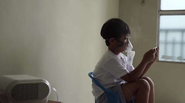 Asian young boy sitting on chair using nebulizer mask with visible mist while looking at smartphone, respiratory treatment at home, pediatric healthcare concept
