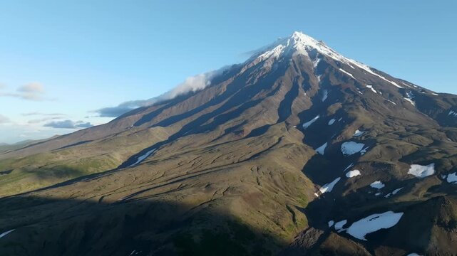 Aerial footage of Koryaksky Volcano with rugged lava slopes, snowfields and drifting clouds in Kamchatka Krai, Russia, dramatic volcanic landscape in clear weather