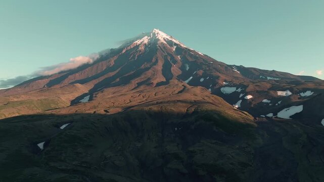 Aerial footage of Koryaksky Volcano with snow patches and volcanic slopes in warm sunrise light, Kamchatka Krai, Russia, with clear sky and clouds