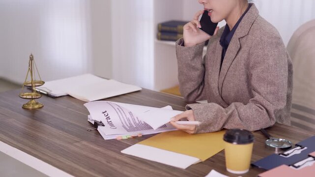 professional Asian lawyer businesswoman talking phone call discussing legal document contract case reviewing paperwork at office desk workspace legal