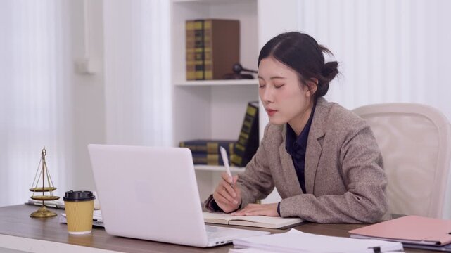 Asian lawyer female attorney review legal document using laptop computer at law office desk with scales justice balance study case research consultation online