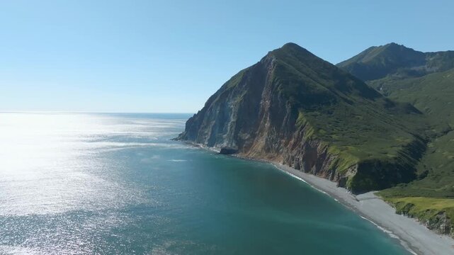 Drone footage of rugged sea cliffs and a quiet pebble beach along Avacha Bay on the Kamchatka Peninsula, Russia, with calm ocean water and clear summer sky