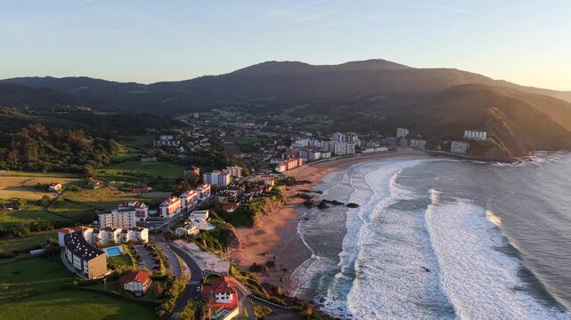 Bakio, Spain drone video - Aerial footage of beach town surrounded by green hills looking out onto the Bay of Biscay near Bilbao at sunset with waves slowly rolling it