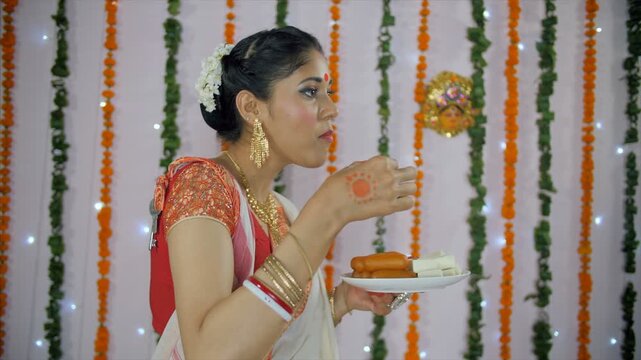 Beautiful Bengali woman eating sweets on Durga Puja festival celebrated in India. Side view shot of a pretty young female enjoying traditional Indian sweets in ethnic saree/sari - festival background
