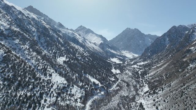 Drone footage of Issyk-Ata Gorge with rugged snow-covered peaks, forested slopes and winding river valley under clear blue sky in Chuy Region, Kyrgyzstan
