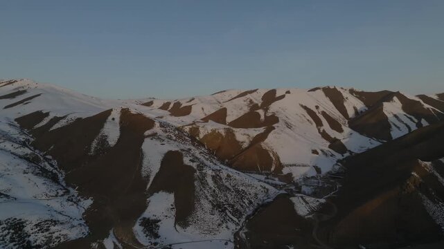 Drone footage of rugged mountain ridges with patchy snow and winding road at golden hour in Chunkurchak Valley, Chuy Region, Kyrgyzstan, Central Asia