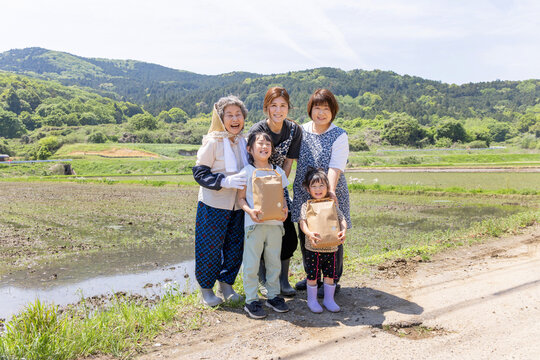 里山の水田で収穫物を手に笑顔を見せる三世代の日本人女性と子供