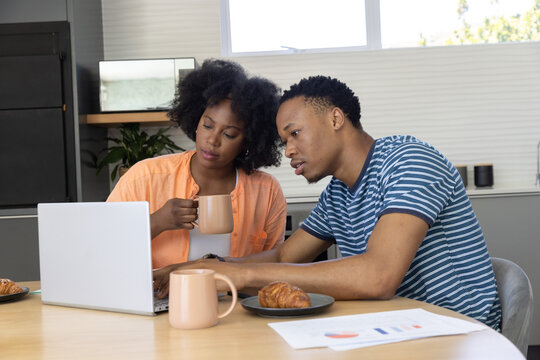 African American couple reviewing charts on silver laptop at wooden dining table holding beige mugs