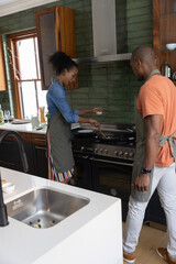 Obraz premium Diverse couple cooking at gas range in kitchen, wearing aprons and stirring food with wooden spoon