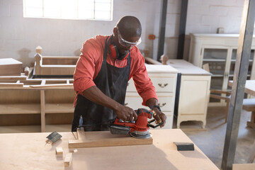 Carpenter sanding wooden plank with red orbital sander at workshop workbench under natural light © wavebreak3