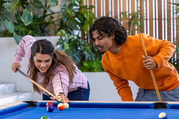 Obraz premium Two friends leaning over blue-felt pool table in plant-filled lounge, lining up shot with cues