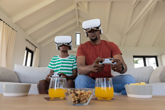 African-American father-son gaming at home wearing VR gear holding pads by table with juice, snack