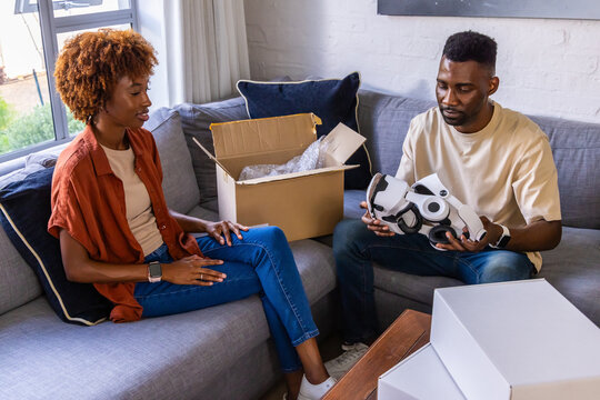 African American couple inspecting white robot on sofa in living room with open box, bubble wrap