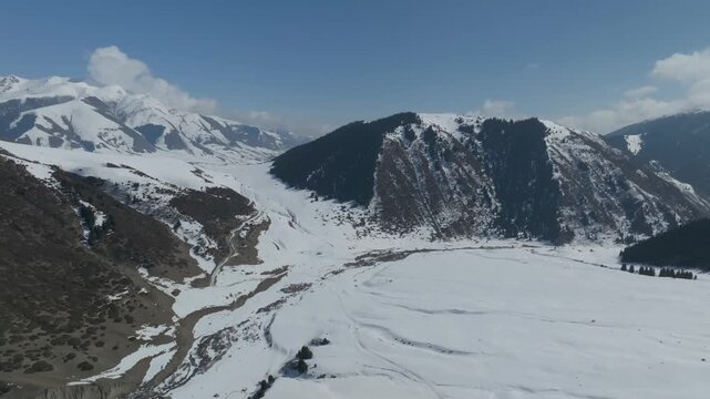 Drone footage of Grigoryev Gorge with snow-covered valley, winding mountain road and forested slopes under blue sky, Issyk-Kul Region, Kyrgyzstan
