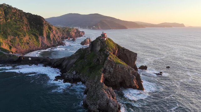 Aerial drone footage orbiting left with wide view of Gaztelugatxeko Hermitage church on rocky islet at sunset on a spring evening, Basque Country Spain