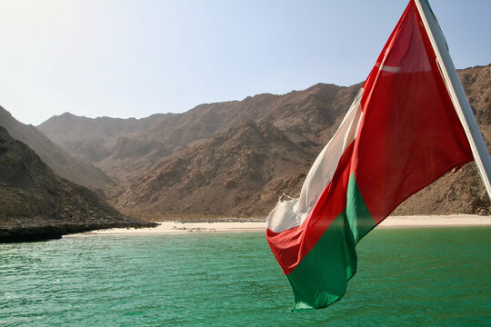 Omani flag flying on dhow boat with turquoise fjord and Hajar Mountains, Musandam, Oman