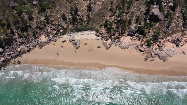 A drone view of turquoise waves crashing on a quiet sandy beach bordered by rocks and forest at Good Ju Ju Lookout, Australia