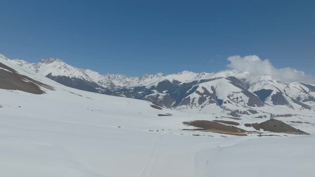 Drone footage of snow covered mountain landscape with a winding road and wide valley under blue sky in Grigoryev Gorge, Issyk Kul Region, Kyrgyzstan