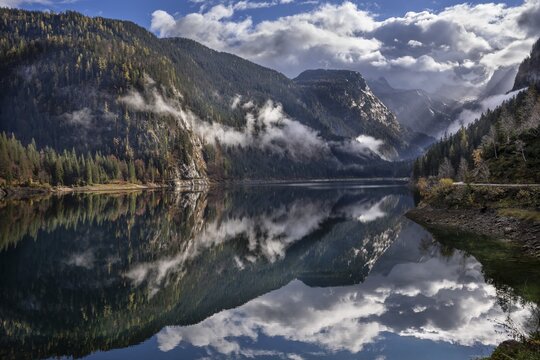The Vordere Gosausee in autumn. In the background, the Dachstein mountain range in clouds. Partly blue sky and sun. Forest covered in clouds. Individual rays of sunshine. Vorderer Gosausee, Gosau, Gosau Valley, Salzkammergut, Upper Austria, Austria