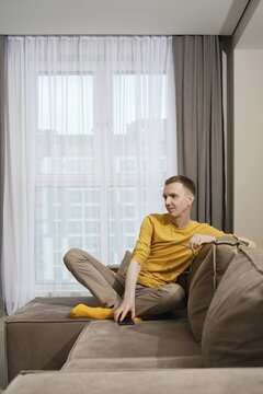 A dreamy man dressed in a yellow shirt and beige pants sits comfortably on a sofa, casually resting his leg in yellow socks