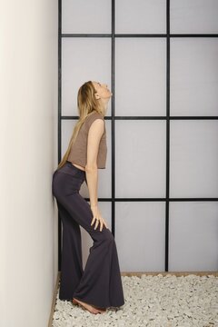 A woman stands in a yoga pose against a wall in a room with a grid design. She wears comfortable clothes and focuses on her balance. The floor has stones, creating a natural look in the space