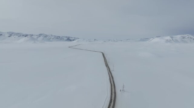 Drone view of a long winding road cutting through the snow-covered Arabel Plateau, Kyrgyzstan, with distant mountain ridges under an overcast winter sky