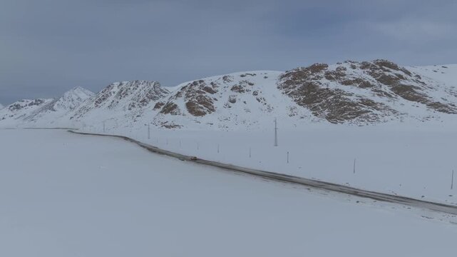 Aerial footage of a remote paved road crossing the snow-covered Arabel Plateau, surrounded by rugged mountains and cloudy winter sky in Issyk-Kul Region, Kyrgyzstan