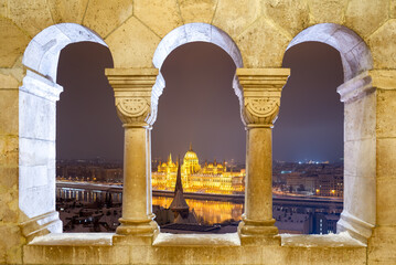 Fototapeta premium Illuminated Hungarian Parliament Building at night framed by triple stone arches of Fisherman's Bastion in winter, Budapest